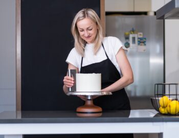 Happy blonde woman finishing processing white mastic on sugar free cake with pastry scraper