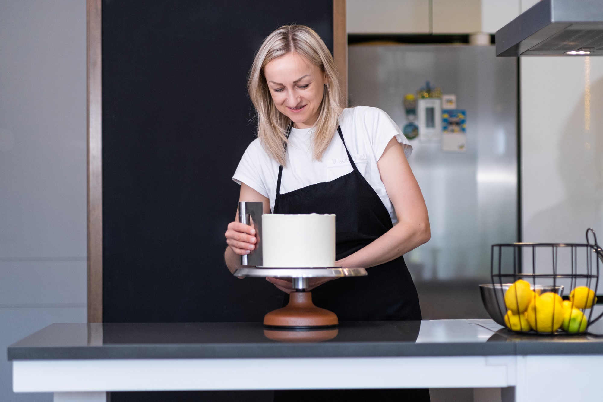 Happy blonde woman finishing processing white mastic on sugar free cake with pastry scraper